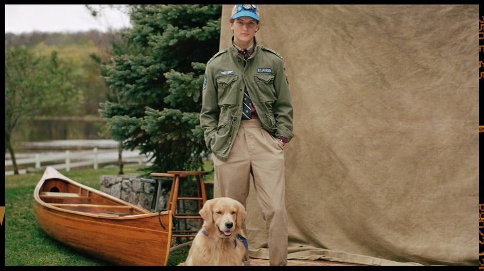 Man in green military jacket & chinos with dog by canoe